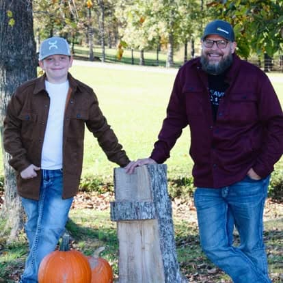 A smiling man and boy pose outdoors next to a tree stump and two pumpkins.