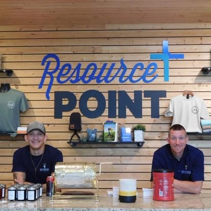Two men smile behind a counter under a Resource POINT sign with a blue cross.