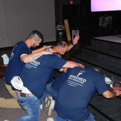 Four men in navy ministry shirts kneel and pray together with hands on shoulders.