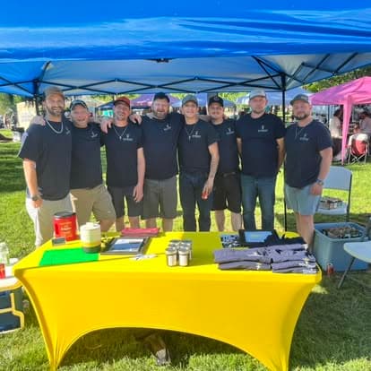 Eight men in dark shirts pose behind a yellow table under a blue canopy.