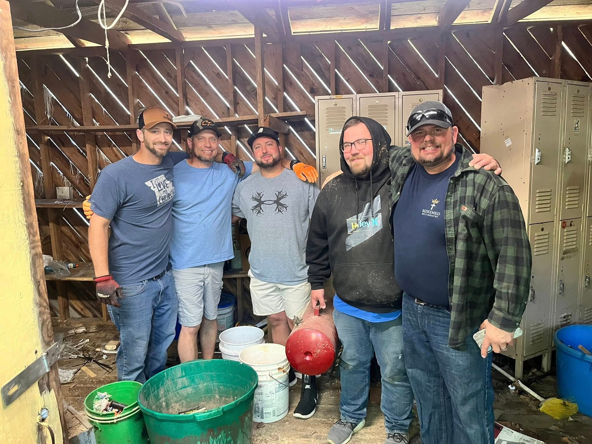 Five men smiling and posing together inside a wooden workshop with lockers and buckets.