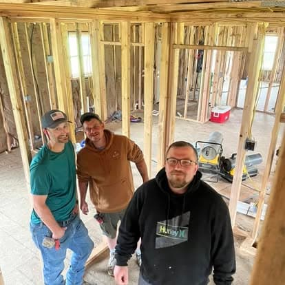 Three men smiling inside a house under construction with exposed wooden wall studs and framing.