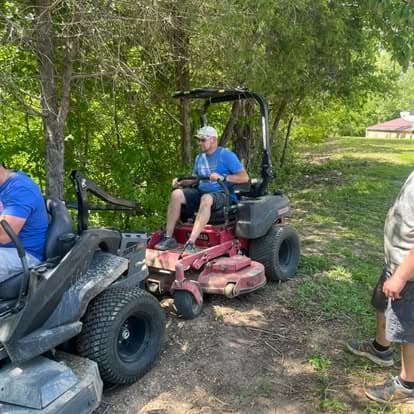 Man in a blue shirt operating a red zero-turn lawnmower near a wooded area.